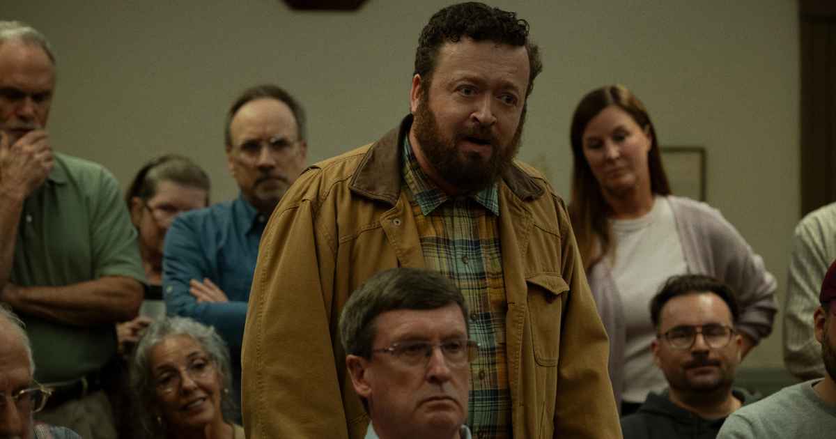 a man in a coat with a beard standing up at a town meeting