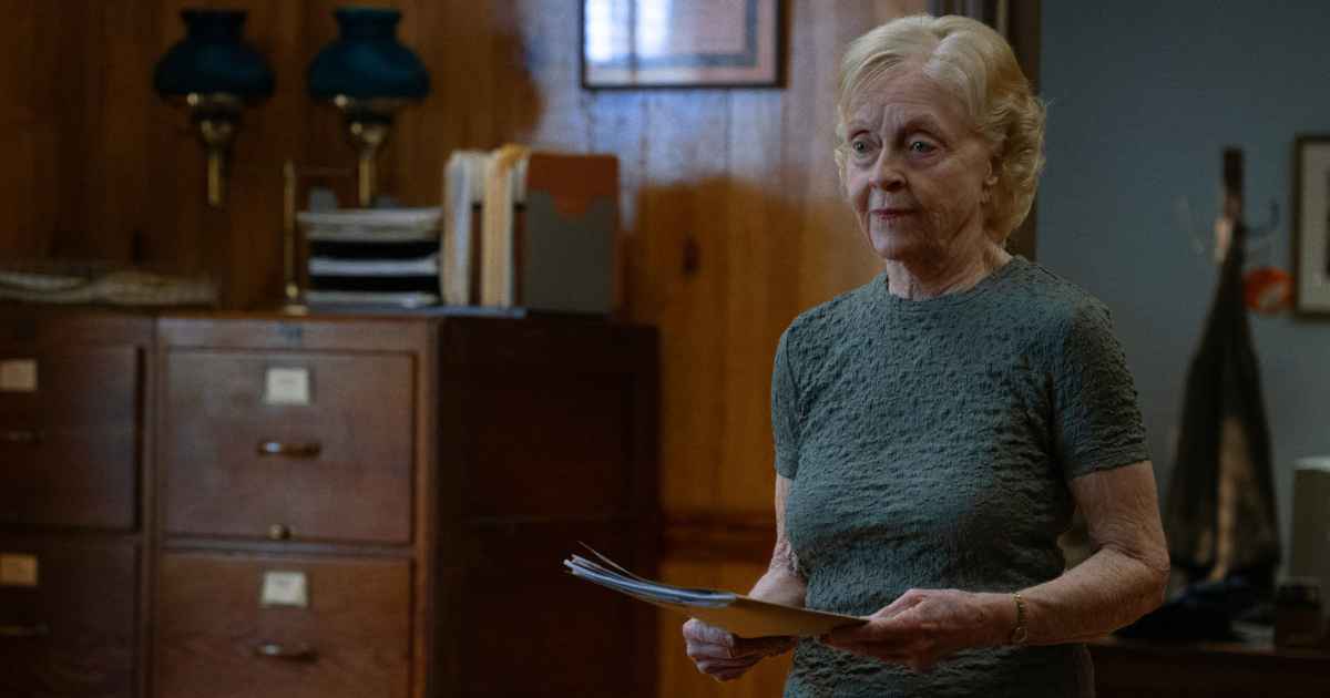 an elderly woman with blue eyes holding a folder standing in an office near a file cabinet