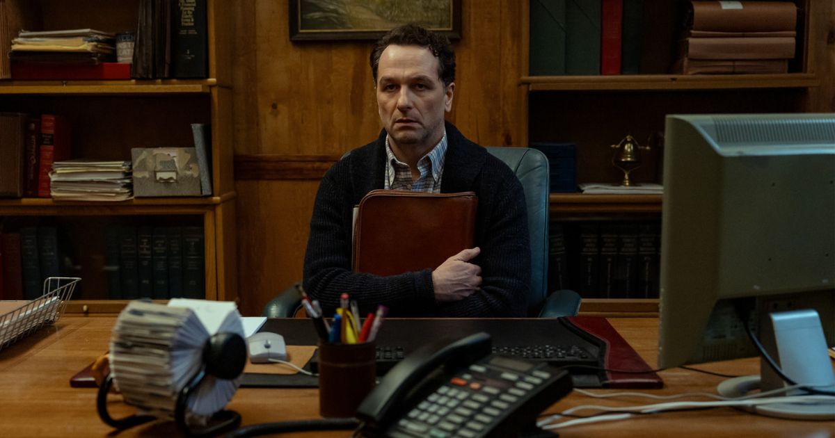 man in blue sweater and plaid shirt sitting at a desk with portfolio looking mortified
