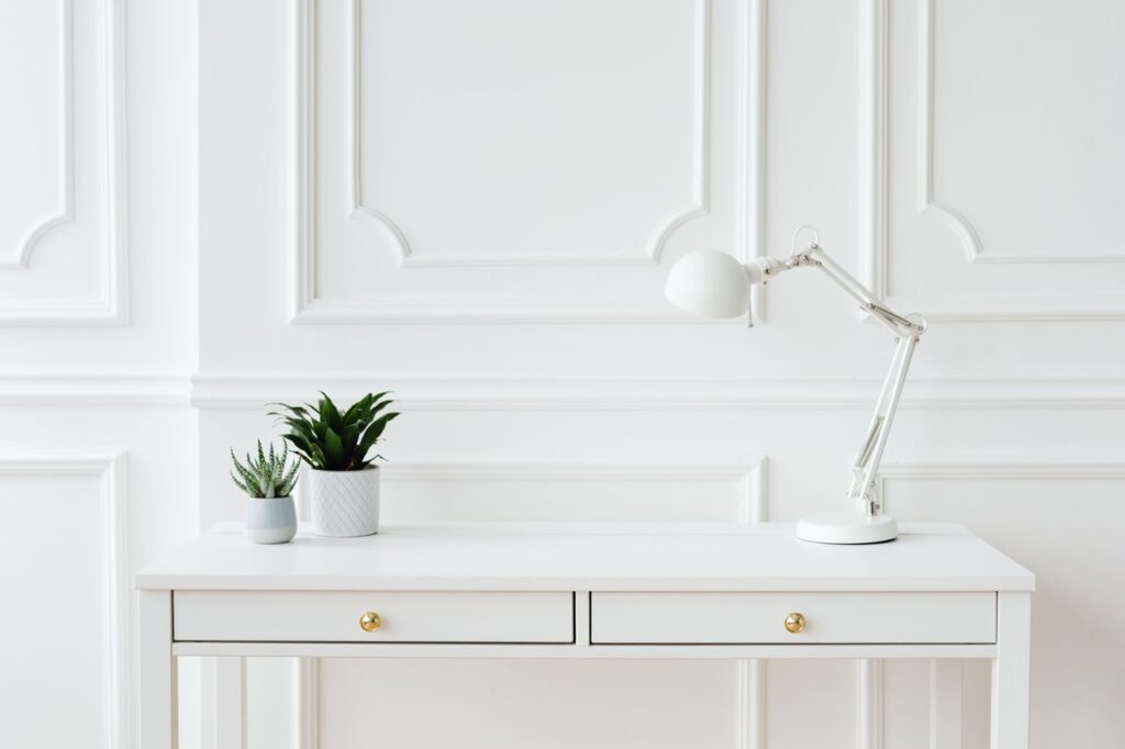 A minimalist white desk setup featuring potted plants and a sleek desk lamp against a white wall.